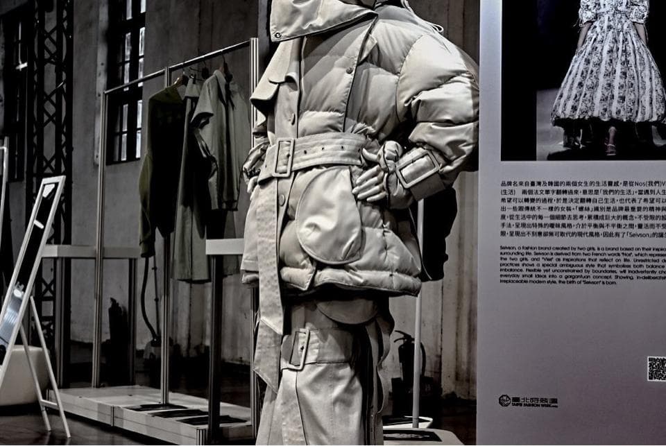 這張「時尚」的照片是一個穿著白色外套的模特,站在一個展示架旁,架子上掛著幾件衣服。背景是水泥牆和一個畫框,畫框中是另一個穿著白色裙子的模特。