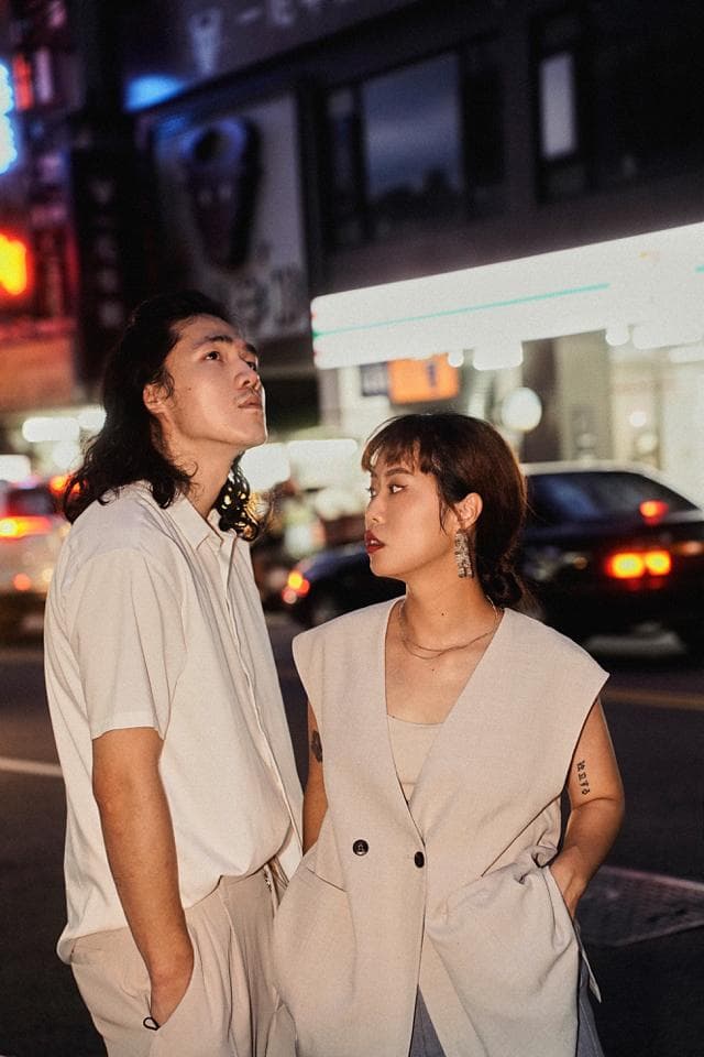 這張照片是兩個年輕人站在一起,他們都穿著淺色衣服。女子穿著米色西裝外套,男子穿著白色襯衫。兩人都在看著遠方,似乎在思考什麼。