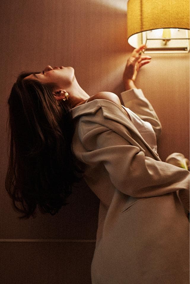 這張「女性側身仰望燈光」的照片是,一位女子身穿白色外套,頭髮披散,頭仰望著天花板上的燈光,眼神充滿著迷離。
