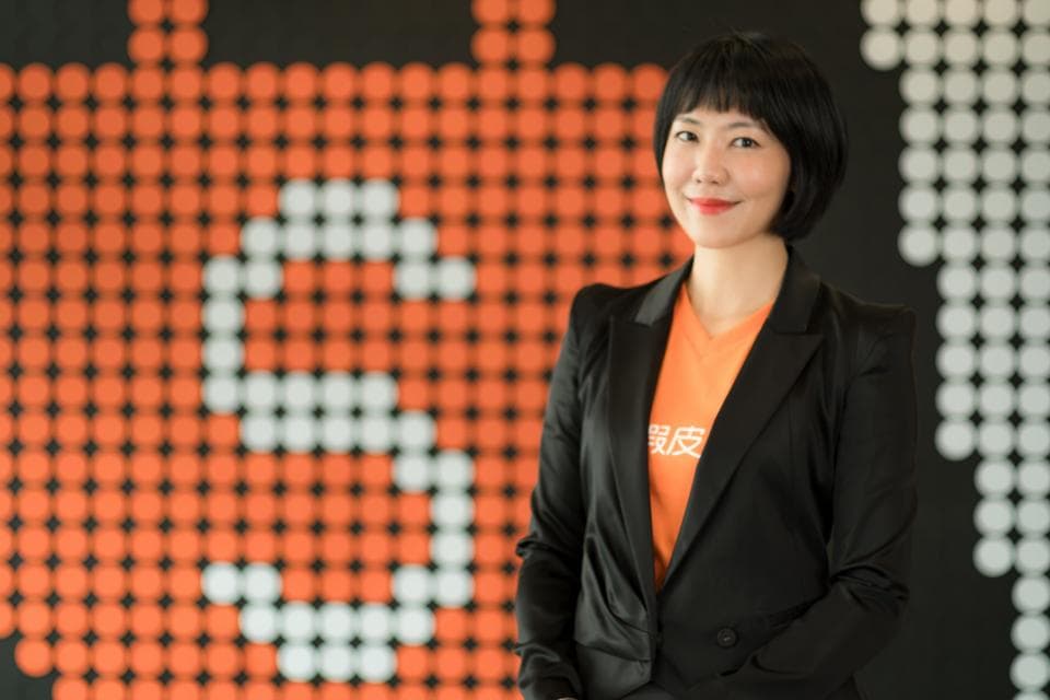 這張「一位女士站在橘色背景前」的照片是。她穿著黑色西裝外套,裡面是橘色T恤,背景上有一個用白色圓點組成的「3」字。