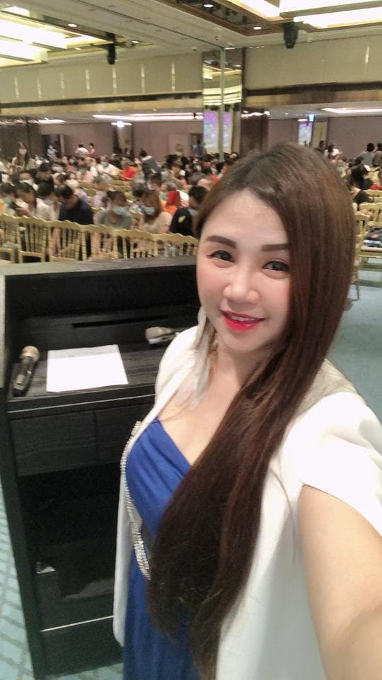 這張照片是一位長髮女士在一個會議室的講臺前,她穿著白色外套和藍色禮服,露出微笑,看著鏡頭。