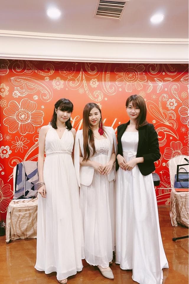 這張「三名女性穿著白色長禮服,站在紅色花紋背景前」的照片是。她們都笑容滿面,看起來很開心。