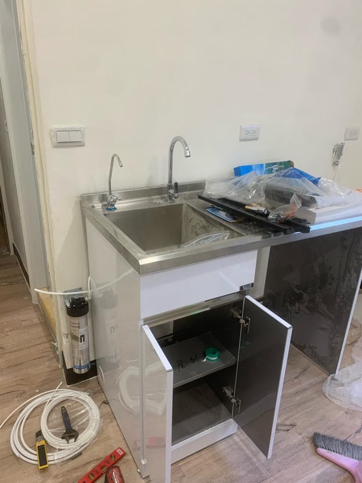 這張照片是一個安裝好的廚房水槽,水槽上有兩個水龍頭,水槽下方是白色的櫃子,櫃子的門已經打開了,露出了裡面的架子。