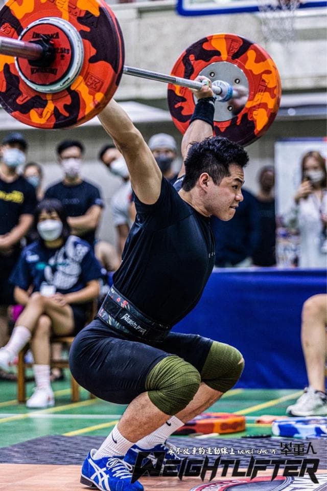 這張照片是一個舉重運動員正在舉起一個槓鈴。他正在用力舉起槓鈴,他臉上流著汗,看起來非常努力。他身穿黑色的舉重服,戴著黑色的舉重手套,腰上戴著一條舉重腰帶。他正在做一個舉重的動作,他正在努力將槓鈴舉起來。