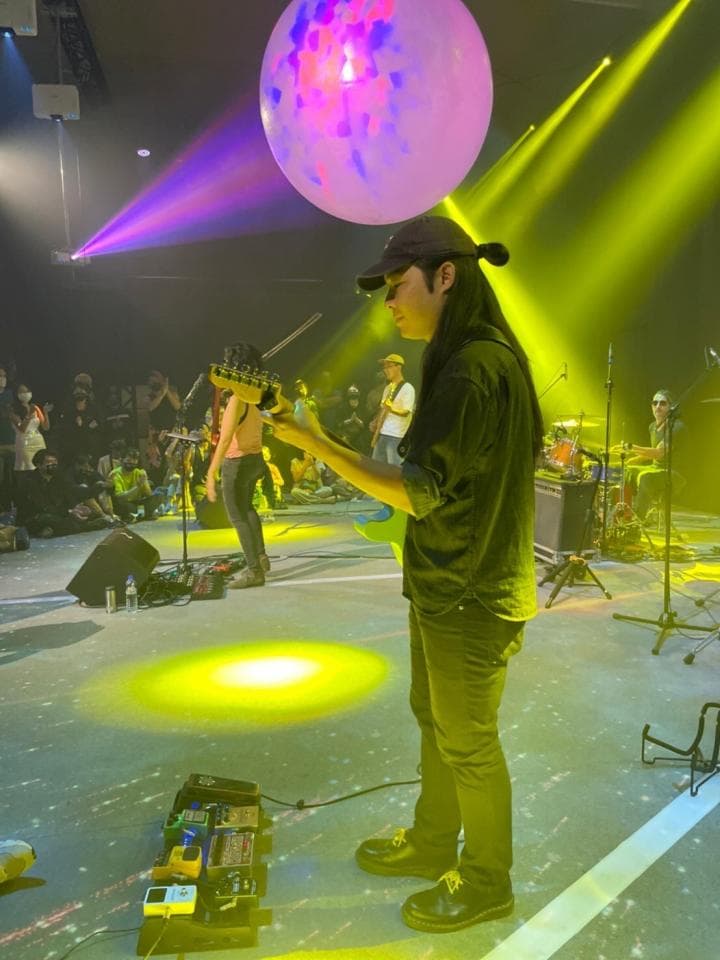 這張照片是一個音樂家在舞台上演奏吉他。他戴著帽子,穿著黑色上衣和牛仔褲。舞台上有一個大型彩色氣球,他正在演奏的吉他上連接了許多效果器。