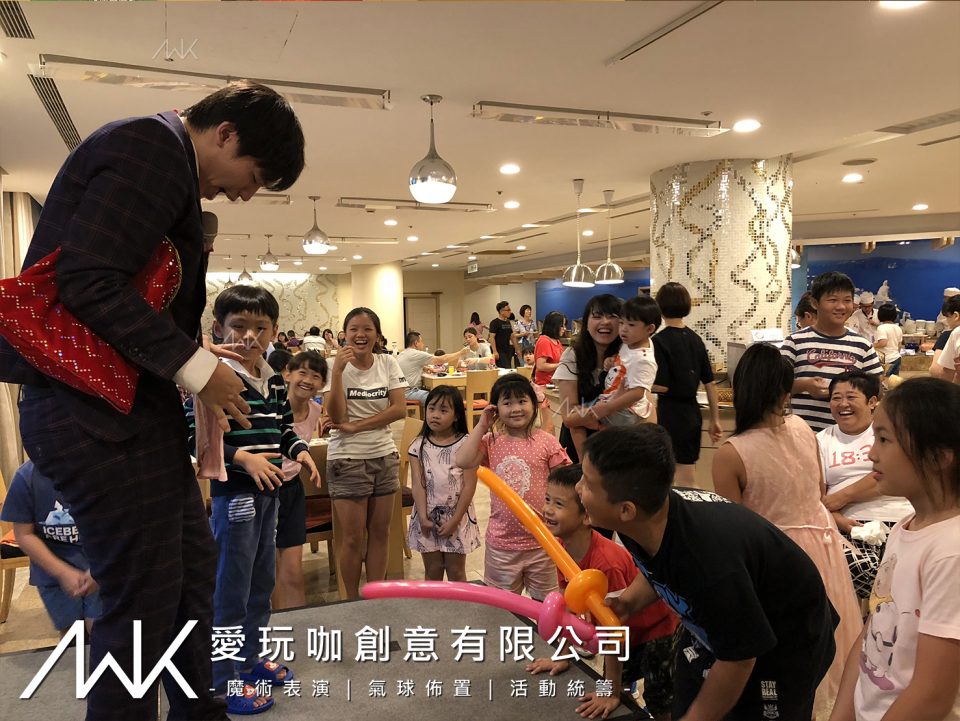 這張照片是一名魔術師正在為一群孩子表演魔術。他身穿西裝,手持一個紅色的魔法袋,正專注地表演。孩子們圍繞在他身旁,目不轉睛地看着他的表演,非常投入。