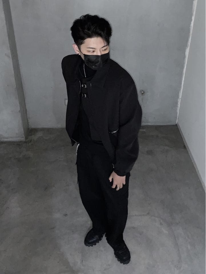 這張照片是一個戴著黑色口罩的年輕男子,穿著全黑的服裝,站在灰色的牆壁前。他穿著一件黑色外套和一條黑色褲子,並配戴一雙黑色的鞋子。