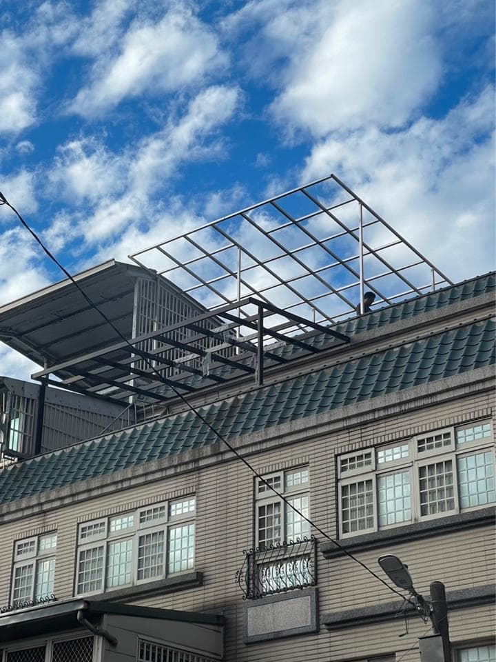 這張照片是一棟有鐵架遮陽的建築物,建築物外牆是米白色,屋頂是深綠色的,屋頂上有一層鐵架,鐵架上有許多小格子,建築物上有許多窗戶,窗戶都採用了百葉窗的設計。