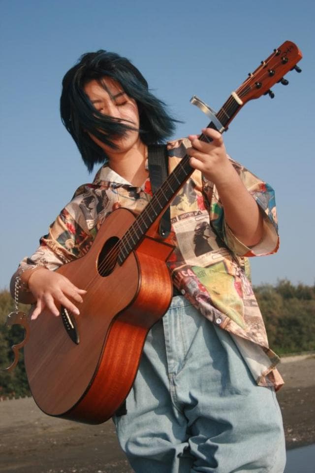這張「一個女生彈吉他」的照片是一個女生在戶外彈奏木吉他,她穿著花襯衫和牛仔褲,背景是藍天。
