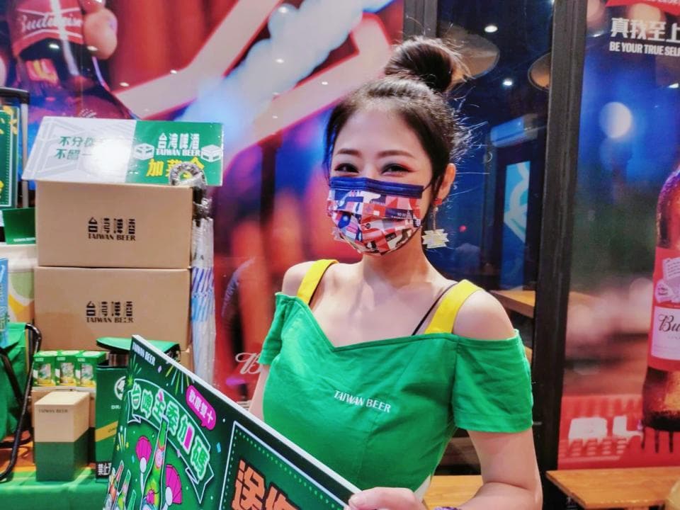 這張「臺灣啤酒」的照片是一個年輕女性,她戴著口罩,穿著綠色的衣服,並拿著一張臺灣啤酒的廣告牌。