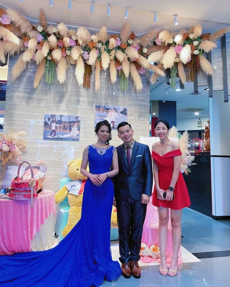 這張婚禮的照片是一個穿著藍色禮服的新娘和穿著紅色禮服的伴娘站在穿著西裝的新郎旁邊,背景是裝飾著乾燥花的花拱門。