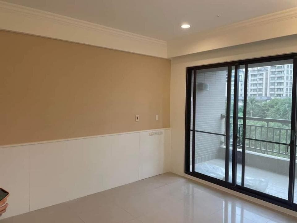 1) 照片顯示一個房間,房間的牆壁是米色的,裝修完成並帶有白色踢腳線,房間裡有落地窗,窗外可以看到建築物和一些樹木