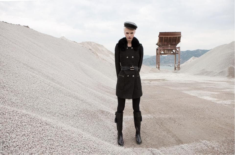 這張「一位年輕女子穿著黑色大衣,站在一片白色碎石灘上的照片」是,照片中女子戴著黑色帽子,並以堅定的眼神凝視前方。她站在一個巨大的沙堆上,周圍是白色碎石,營造出一個荒涼而神秘的氛圍。