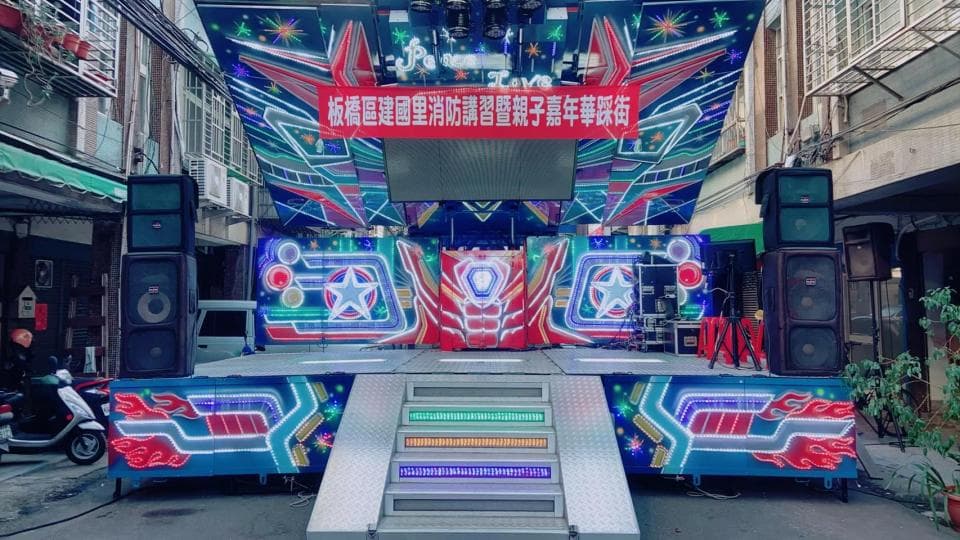 1) 這是一張在台灣街道上拍攝的照片,照片中有一個裝飾有LED燈的舞台,上面有一塊紅色的布條,上面寫著「歡迎來到這裡」