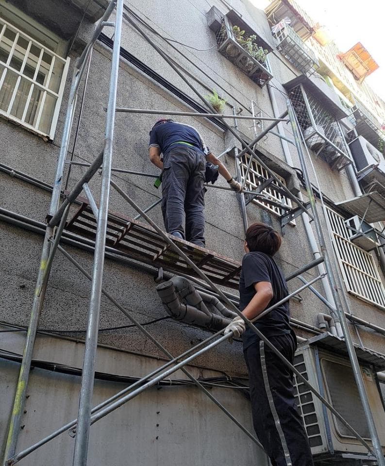這張照片是兩個工人站在鷹架上,在建築物外牆進行維修工作。