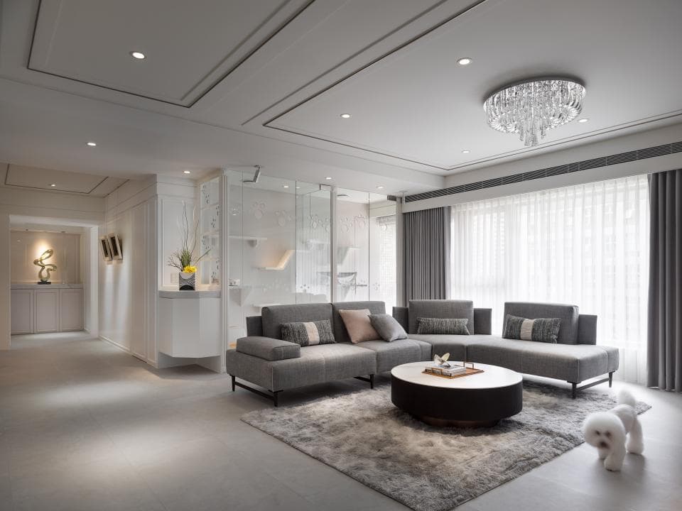 這張「現代風格客廳」的照片是,以白色和灰色為主色調,有灰色布沙發、圓形咖啡桌、地毯和白色的小狗。牆壁上的裝飾品,以及天花板上的水晶燈,都增添了空間的質感。