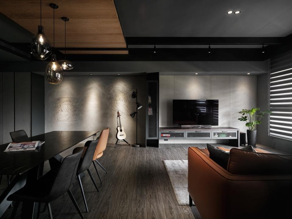 這張「現代風格的客廳」的照片是一間以灰色和黑色為主色調的空間,中間擺放了一張黑色長桌,搭配幾張灰色和棕色椅子,左邊牆上掛著一架木吉他,右邊牆上有一個大型電視,沙發則選擇了橘棕色,整體空間營造出沉穩的氛圍。