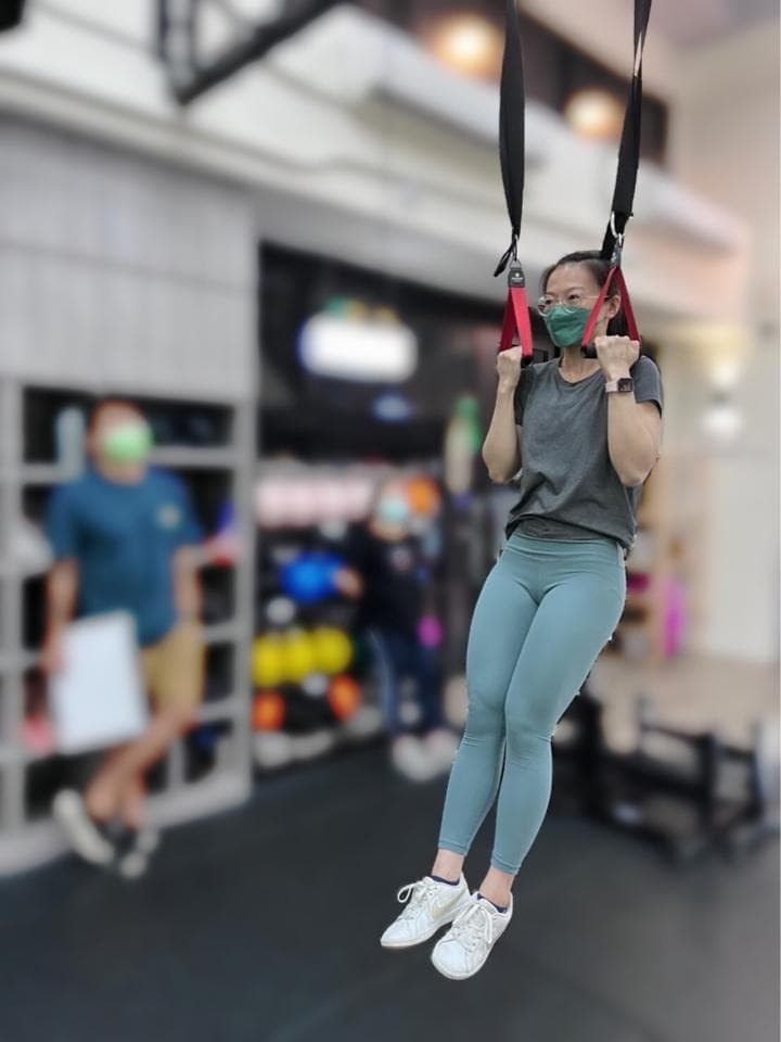 這張照片是一個穿著藍色緊身褲的女性,她正在健身房做懸吊訓練。她戴著一個灰色的口罩,用手抓住懸吊繩索,看起來很專注。