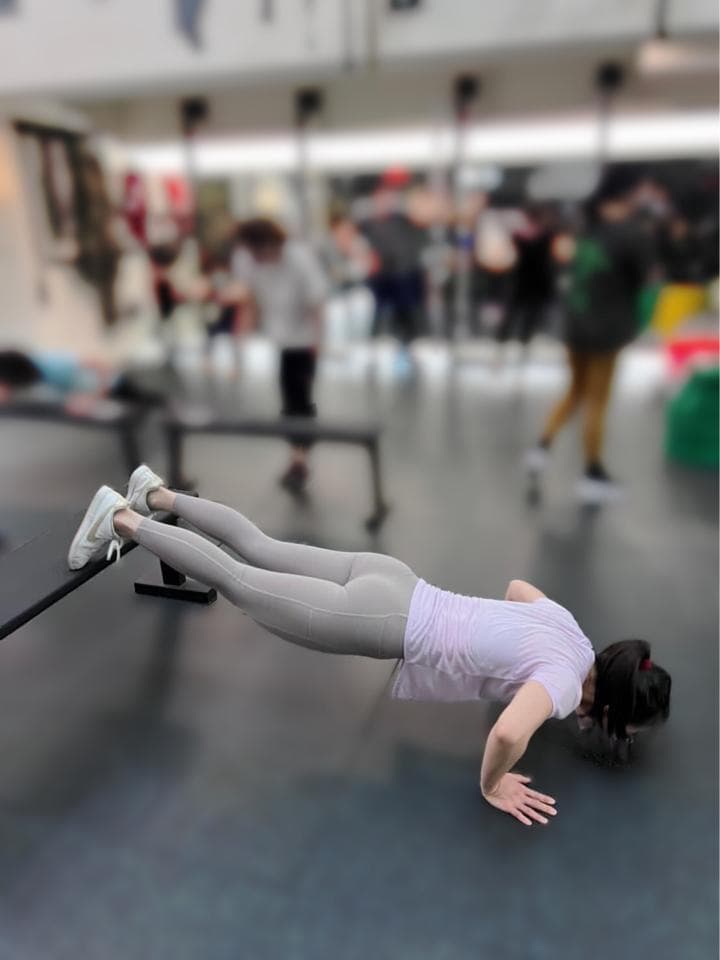 這張照片是一名女子在健身房做伏地挺身,她穿著灰色的緊身褲和白色的運動上衣,雙手撐著地板,身體呈現一條直線,姿勢非常標準。