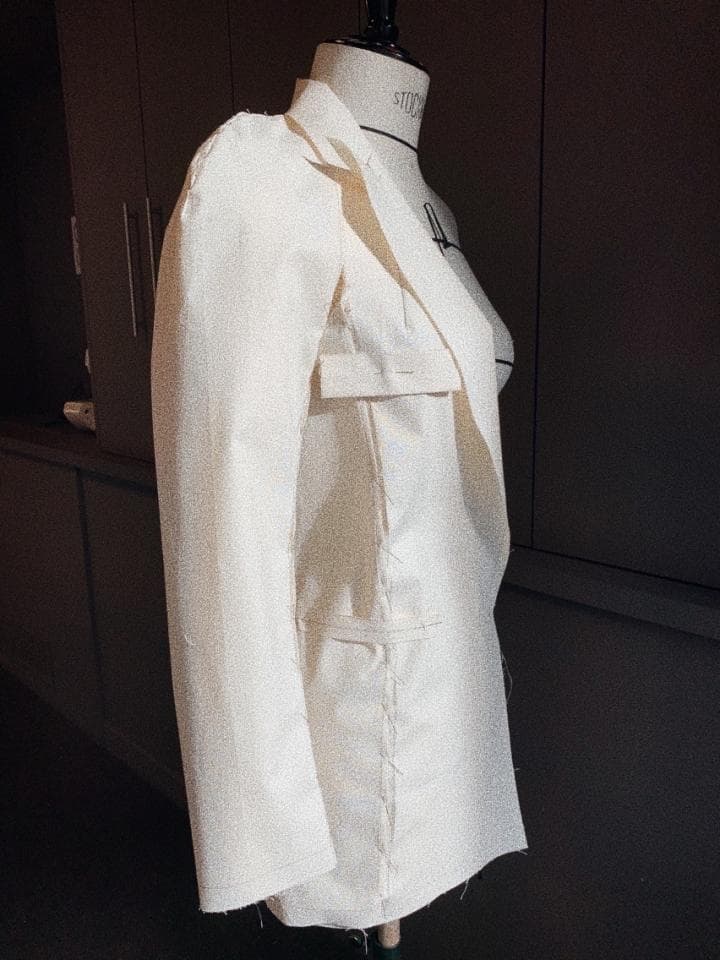 這張照片是一件白色的西裝外套,它被放置在一個裁縫模型上。外套的縫線細節清晰可見,突顯了裁縫的精湛技藝。