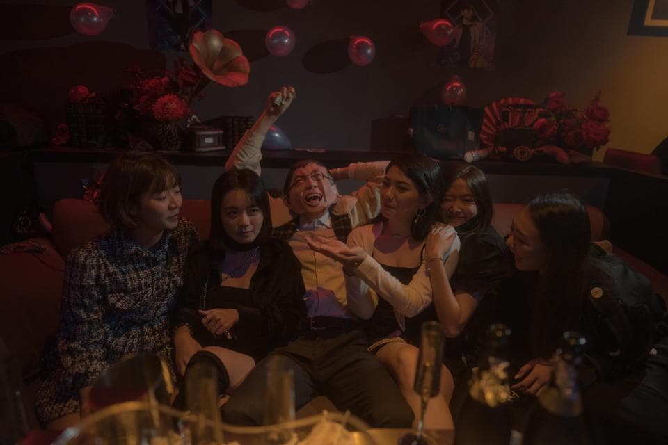 這張照片是一群朋友在室內聚會,他們圍坐在沙發上聊天,看起來很開心。照片的中心是兩位年輕女子,她們看起來非常親密,像是情侶。她們的笑容和眼神充滿愛意,她們的互動是照片中最吸引人的部分。