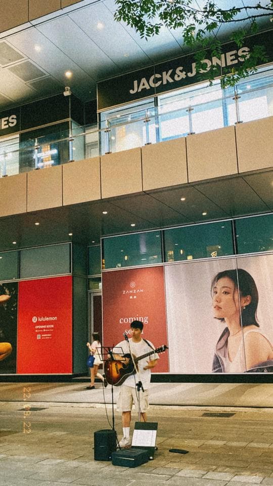 這張照片是一名男子在街頭表演。他戴著帽子,穿著白色短袖上衣和牛仔褲,正在彈著一把木吉他。他面前放著一個音箱和一個話筒。他正在街頭表演,周圍圍觀著一些路人。