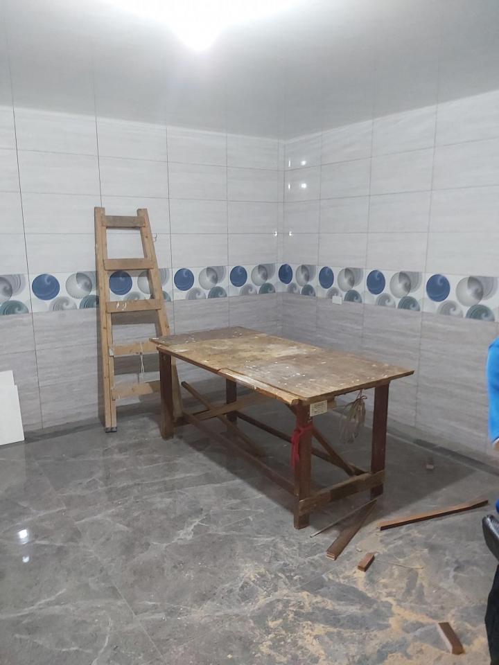 1) 照片顯示一個空房間,房間裡有一張木桌和一個梯子,房間的牆壁上貼著白色和藍色的磁磚,地板是灰色的