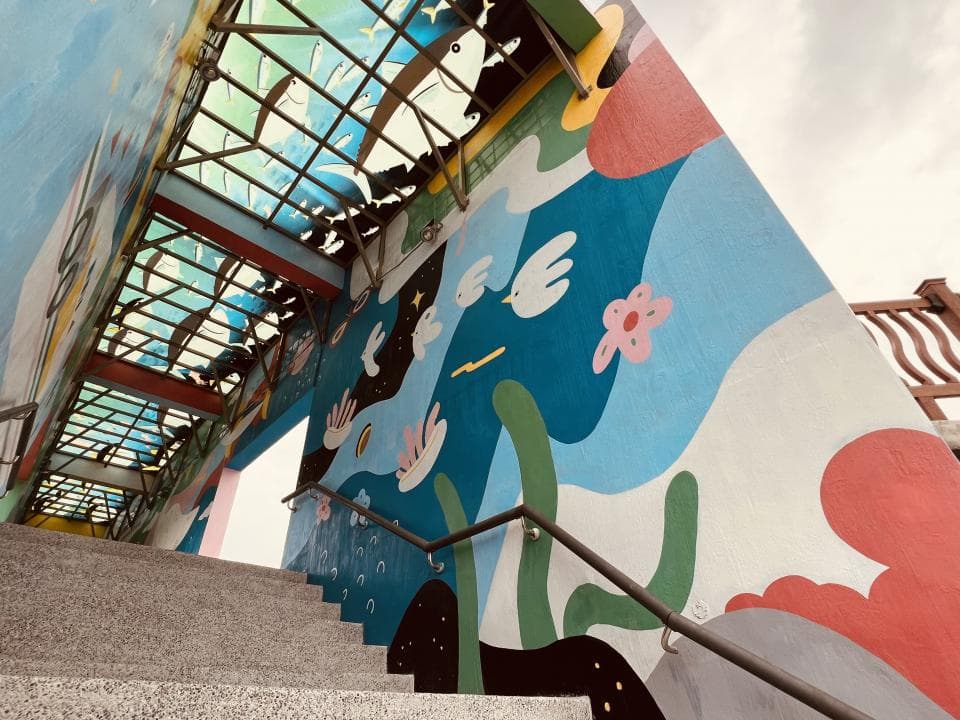 這張「塗鴉牆」的照片是一條有樓梯的通道,牆上畫著藍色、綠色和粉紅色的圖案,還有一些鳥類和植物的圖案。