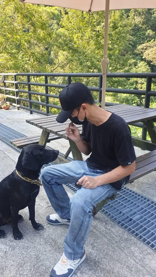 這張「一個男人坐在戶外椅子上,他穿著黑色的T恤和牛仔褲,戴著黑色的帽子,一隻黑色的狗坐在他旁邊」的照片是。