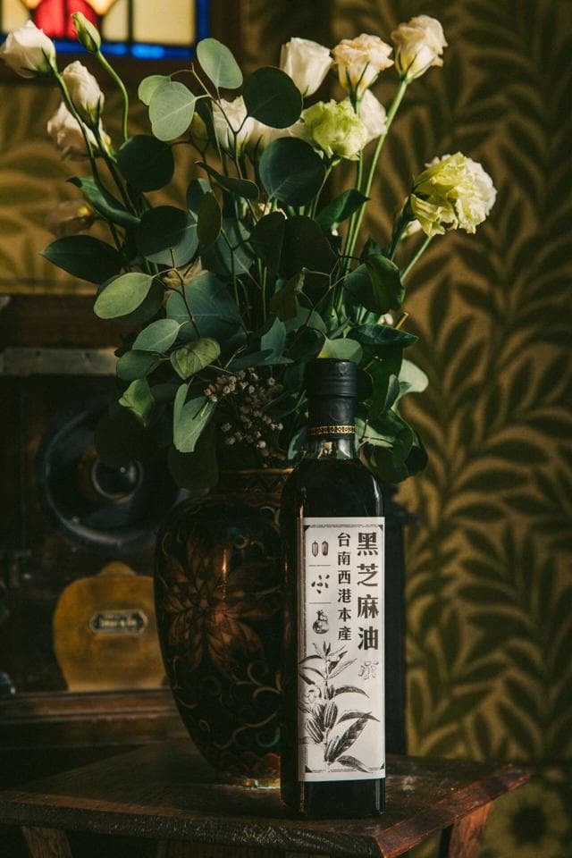 這張照片是一瓶黑芝麻油,瓶身印有產品名稱及成分說明。瓶子放在一個木製桌面上,旁邊有一束白色花朵。
