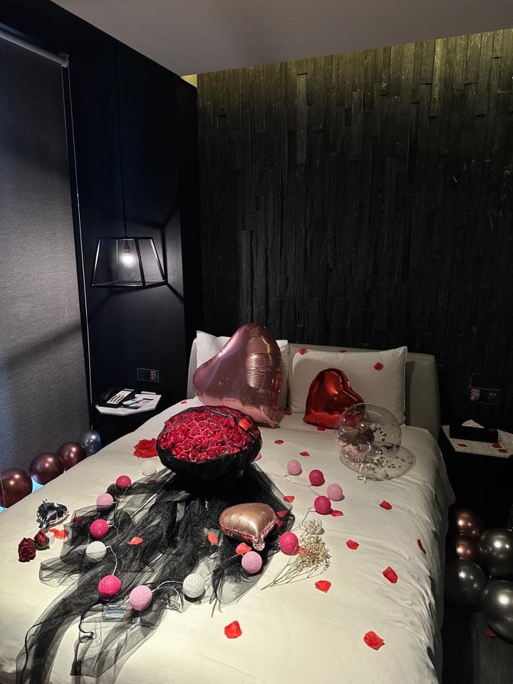 這張「浪漫房間佈置」的照片是一張床,床上鋪著白色床單,上面擺放著花束、氣球和一些裝飾品。床頭板是黑色的木板,牆壁是黑色的。