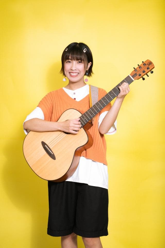 這張照片是一個年輕女性在黃色背景下彈吉他的照片。她穿著橘色的毛衣和黑色的短褲,笑容燦爛。最重要的是她手中的吉他,它看起來很新,而且她彈奏的姿勢也很專業。