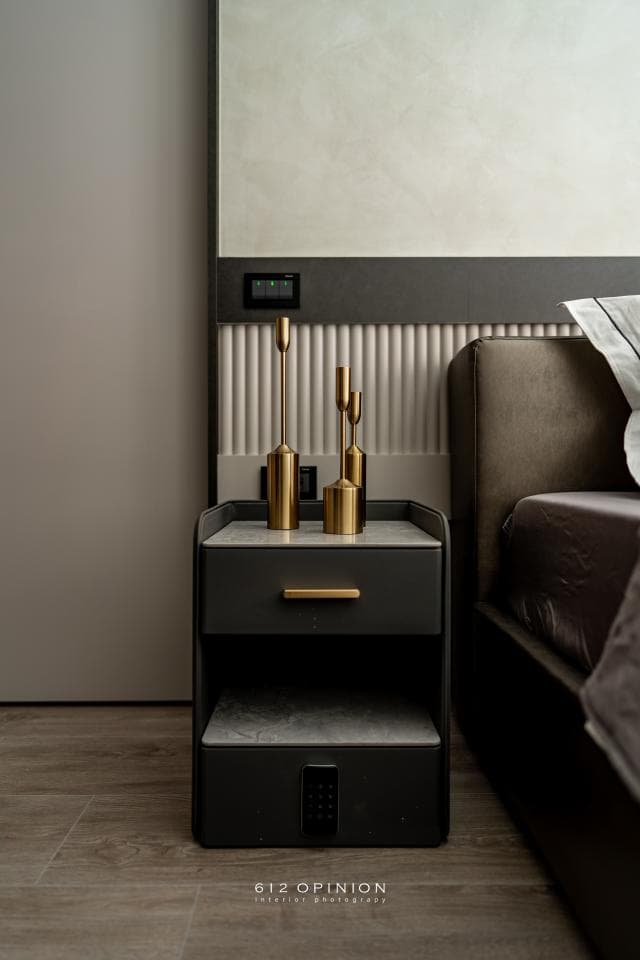 這張「床頭櫃」的照片是一個黑色床頭櫃,上面放著金色的燭臺,床頭櫃旁邊是一張床,床頭是灰色的牆壁。