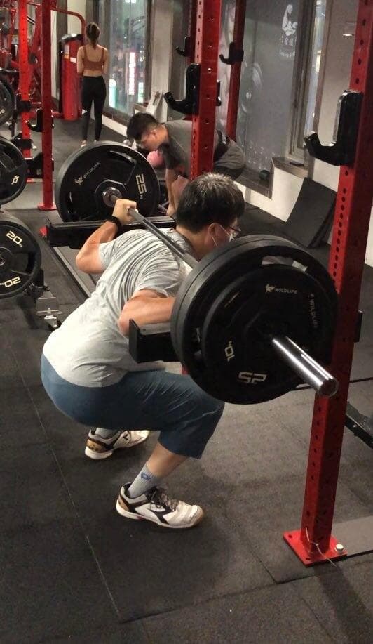 這張照片是一名男子在健身房舉重,他正在做深蹲,身上穿著灰色的衣服,手上舉著一個重重的槓鈴。