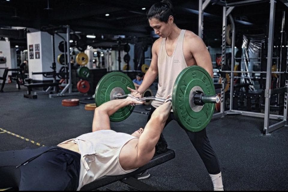 這張照片是一個健身房,照片中最重要的部分是兩個男人在做臥推。其中一個男人正在臥推,另一個男人在後面扶著他的腳,以確保他安全。他們兩個都專注於訓練,表現出他們對健身的熱情。