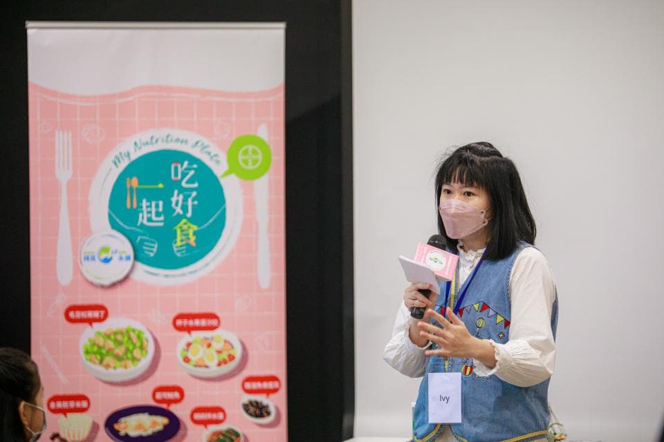 這張照片是一名戴著粉紅色口罩的女生,她手持麥克風,站在一個寫著“吃得好”的宣傳板前面,並且正準備講話。