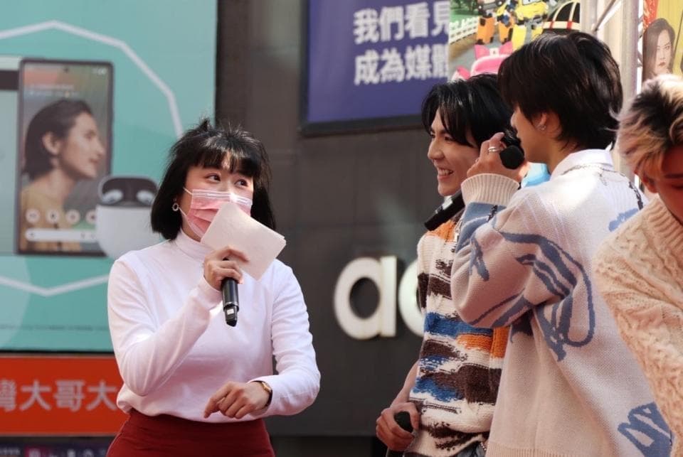 這張「宣傳活動」的照片是一個戴著口罩的女性主持人,她拿著麥克風,正在對著鏡頭說話,在她旁邊有三個年輕人,他們看起來很開心,可能是主持人活動的嘉賓。