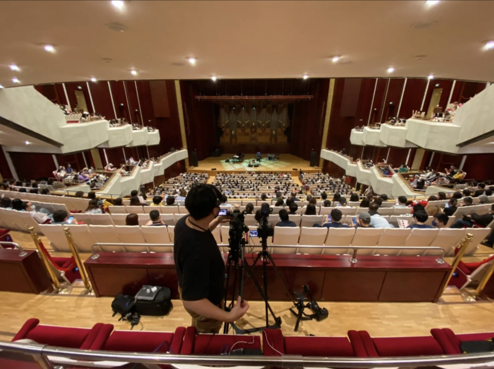 這張「音樂廳」的照片是,一個攝影師站在觀眾席上,使用攝影機拍攝臺上的音樂表演。