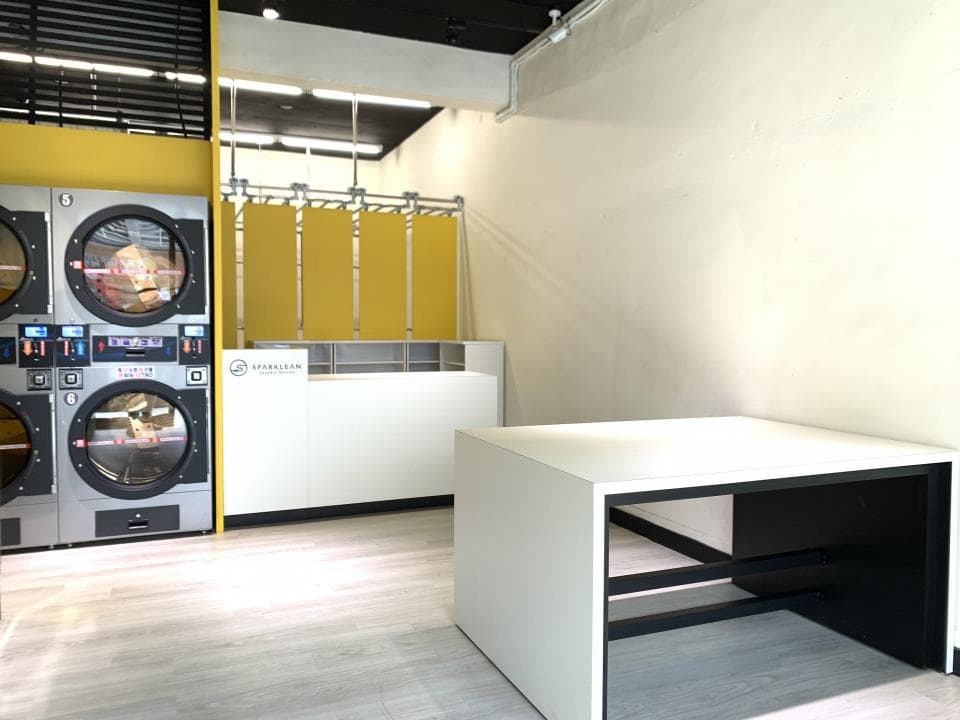 1) 照片顯示一個洗衣店的內部,左邊有兩台大型洗衣機,右邊有櫃台和一個白色桌子