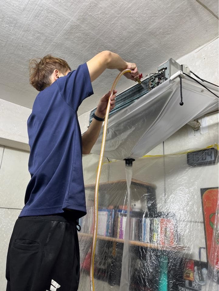 1) 照片顯示一位身穿藍色上衣和黑色長褲的男性正在修理冷氣,他用一根黃色的軟管將冷氣的冷媒排放到一個透明的塑膠袋裡,冷媒從塑膠袋裡流出來,照片的背景是牆壁和書架