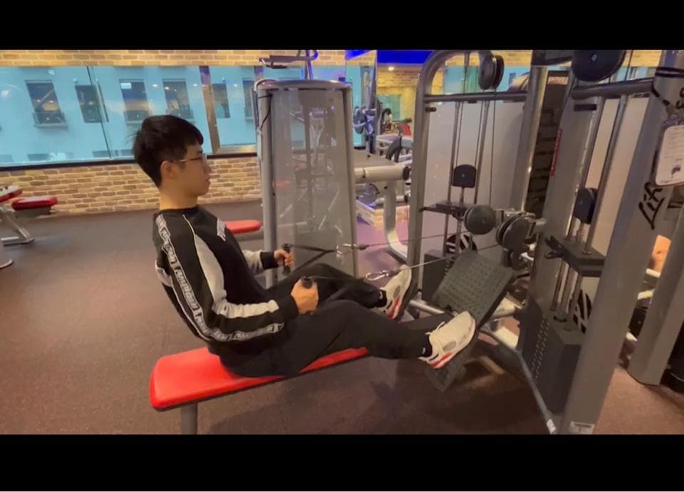 這張照片是一個健身房,照片中的重點是一個年輕人在用拉力器訓練。他穿著黑色運動服,看起來正在努力地拉著重量,他的表情專注而堅定。