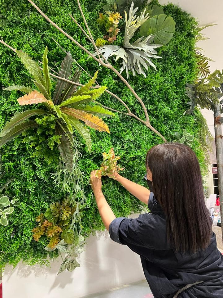 1. 這張照片顯示一個女人站在一面綠色的植物牆前面,牆上有各種不同的植物,包括蕨類植物、綠色和黃色植物,以及一些裝飾性植物