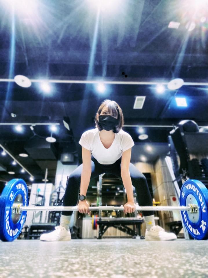 這張照片是一個女生在健身房舉重,她戴著口罩,專注地舉起槓鈴。