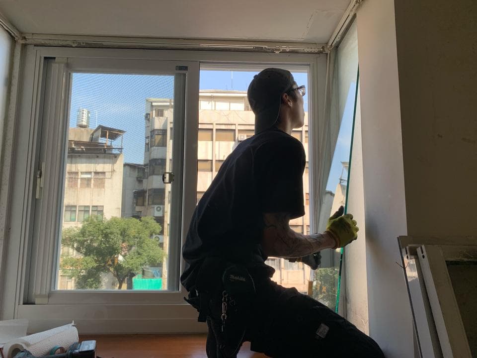 1) 照片顯示一名身穿黑色T恤和工作褲的男子,他站在一個窗戶前,手裡拿著一塊玻璃,準備安裝