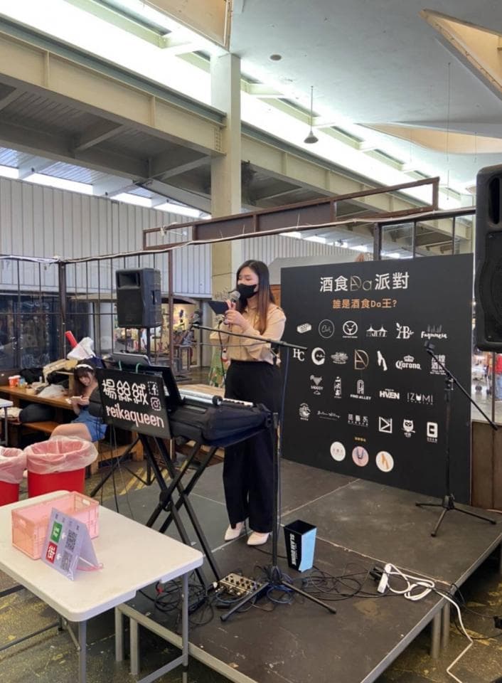 這張「街頭表演」的照片是一位女生在舞臺上唱歌,她穿著黑色衣服,戴著口罩,手持麥克風,並有一臺電子琴。舞臺上有黑色的佈景,上面印著許多品牌標誌。