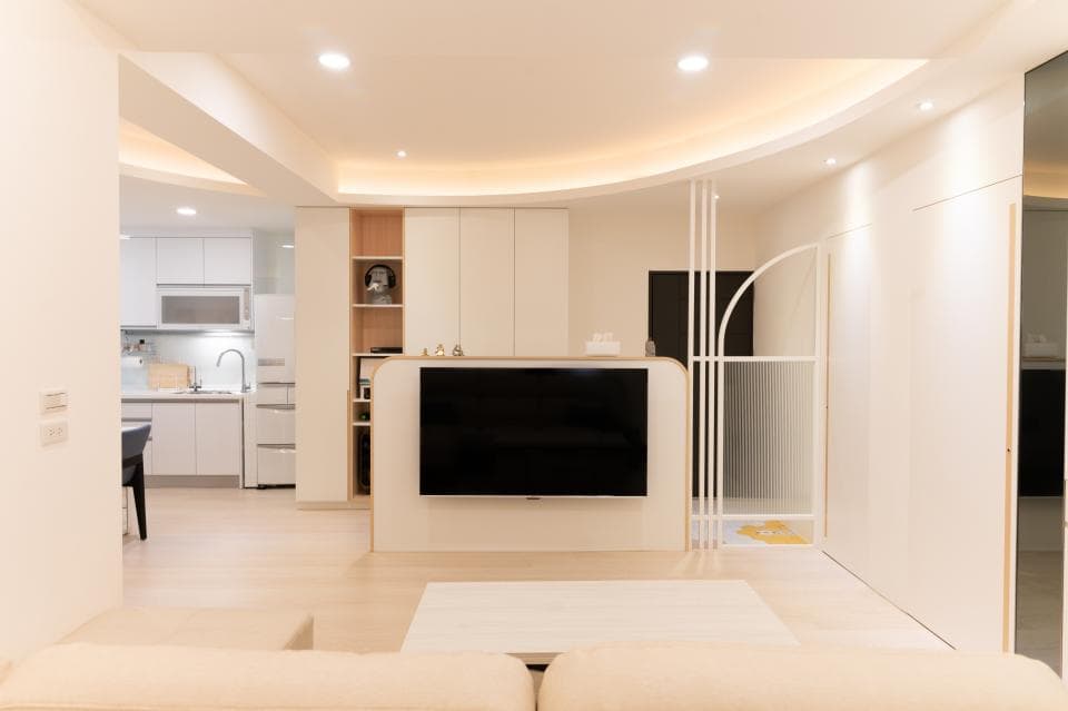 這張「現代化客廳」的照片是一間明亮且簡潔的客廳,擁有大型電視、白色牆壁和淺色木地板。房間的左側是一個開放式廚房,可以看見流理臺和冰箱。右側則是一個以白色格柵隔開的房間。