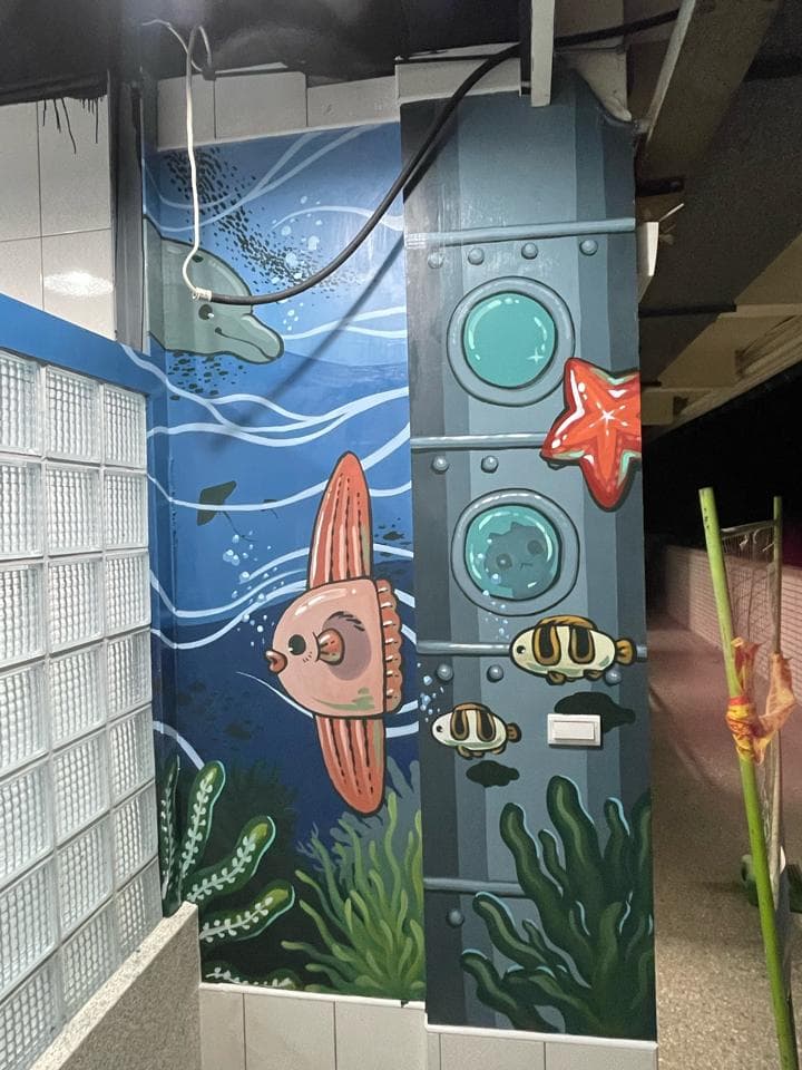 這張照片是一幅牆上繪製的海底世界壁畫,畫面左側是海豚和各種海洋生物,右側是一艘潛水艇,潛水艇上有三個圓形窗戶,壁畫的色彩鮮豔,充滿了童趣。