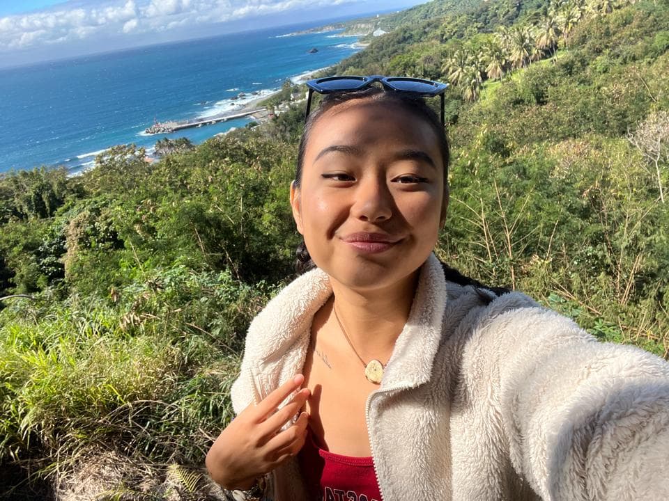 這張照片是一個年輕女性站在山坡上,背後是一片碧藍的海。她戴著墨鏡,穿著白色毛衣,笑容滿面。