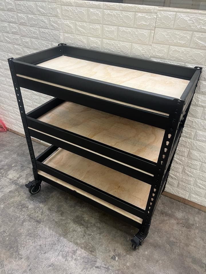 1) 照片顯示一個黑色的金屬架子,有三個木板層架,架子有輪子,架子放在牆壁旁的地板上