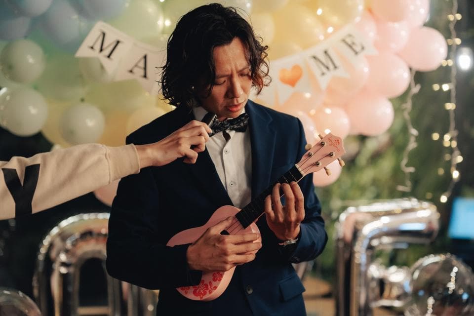 這張照片是一個男人彈著一把粉紅色的尤克里裡,他穿著黑色的西裝外套和白色的襯衫。